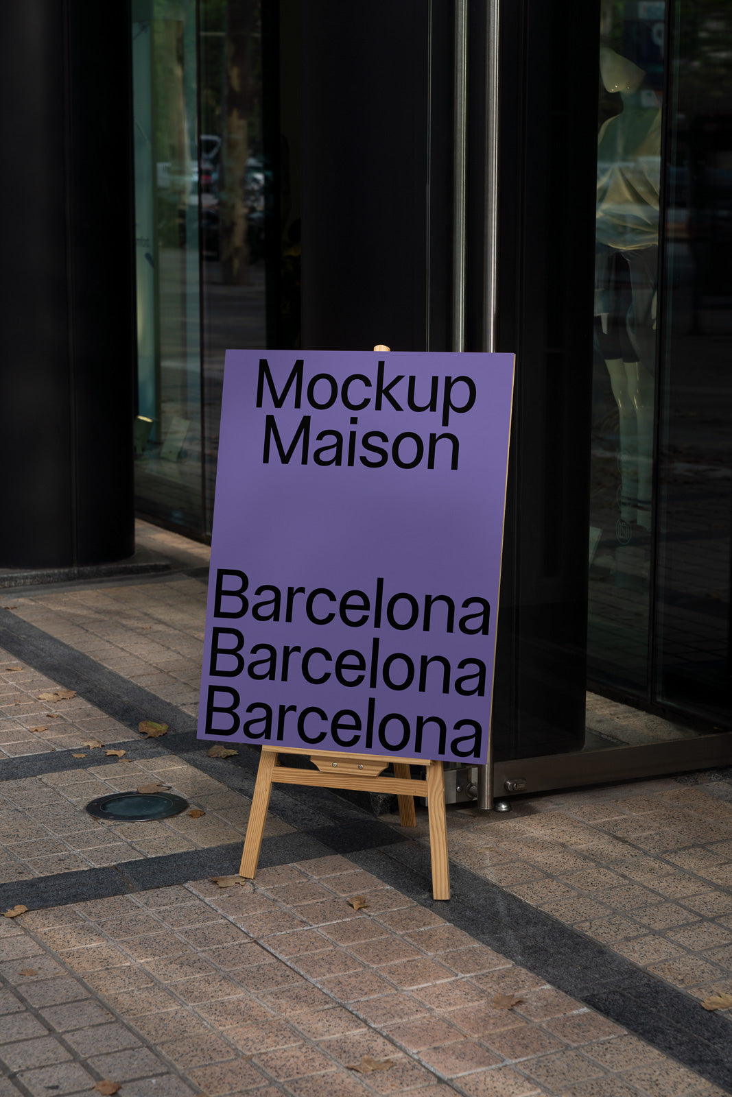 PSD mockup of a purple poster on an easel with black text reading "Mockup Maison Barcelona" in front of a glass building entrance.
