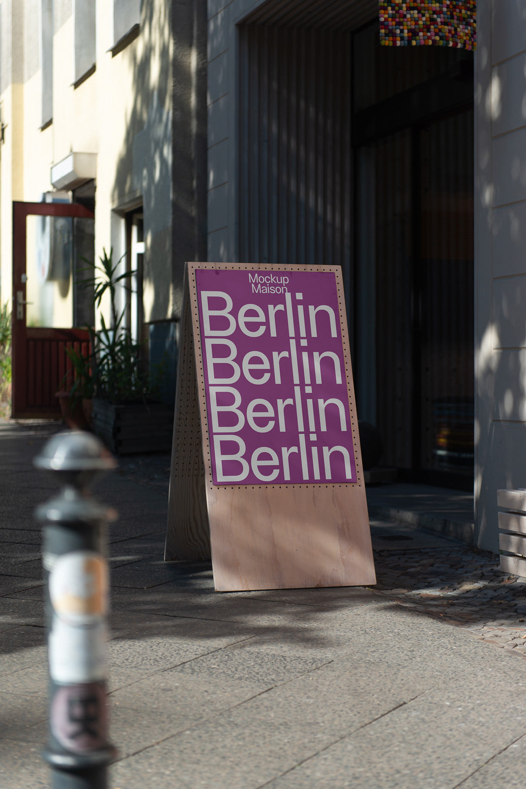 PSD mockup of a wooden stand on a city sidewalk displaying a sign with the word "Berlin" repeated on a purple background.