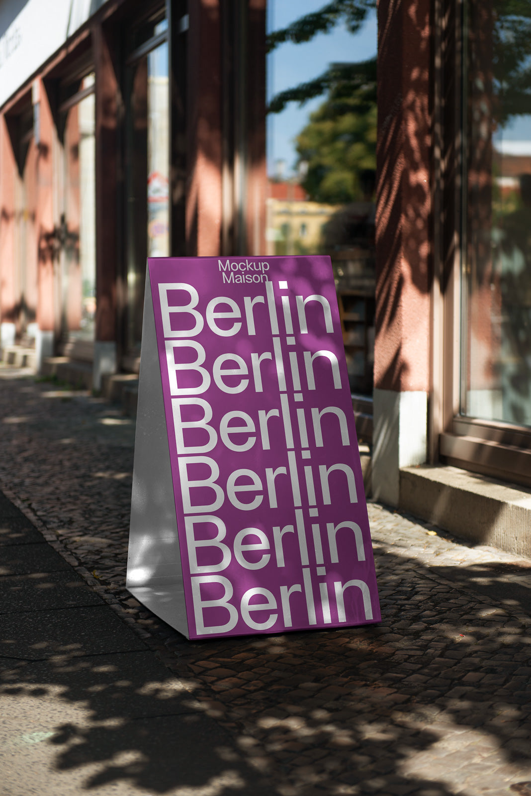PSD mockup of a sidewalk A-frame sign with "Berlin" repeated in white text on a purple background, placed in a sunlit urban setting.