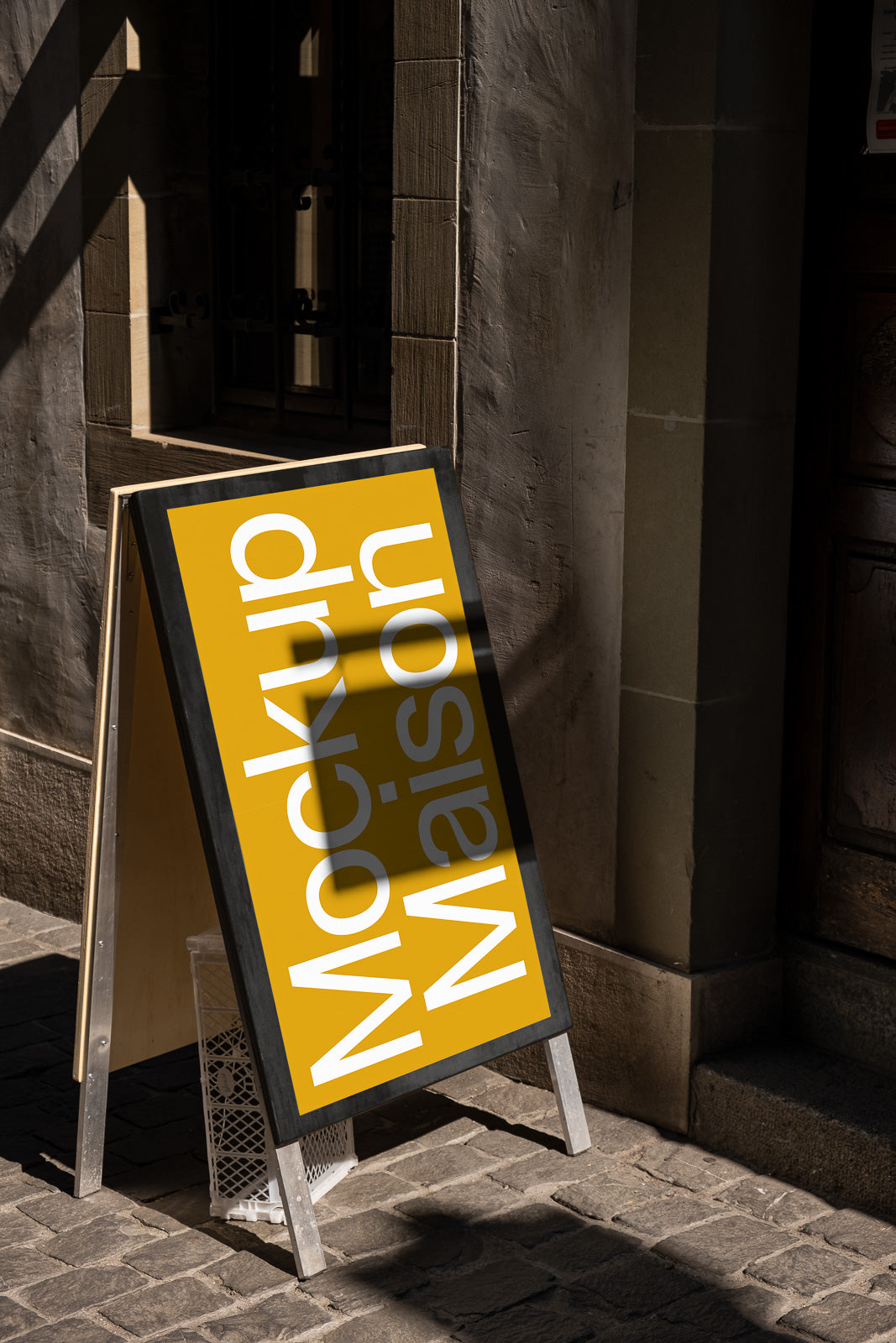 PSD mockup of a yellow sandwich board with white text reading "Mockup Maison" displayed on a cobblestone street next to a textured stone wall.