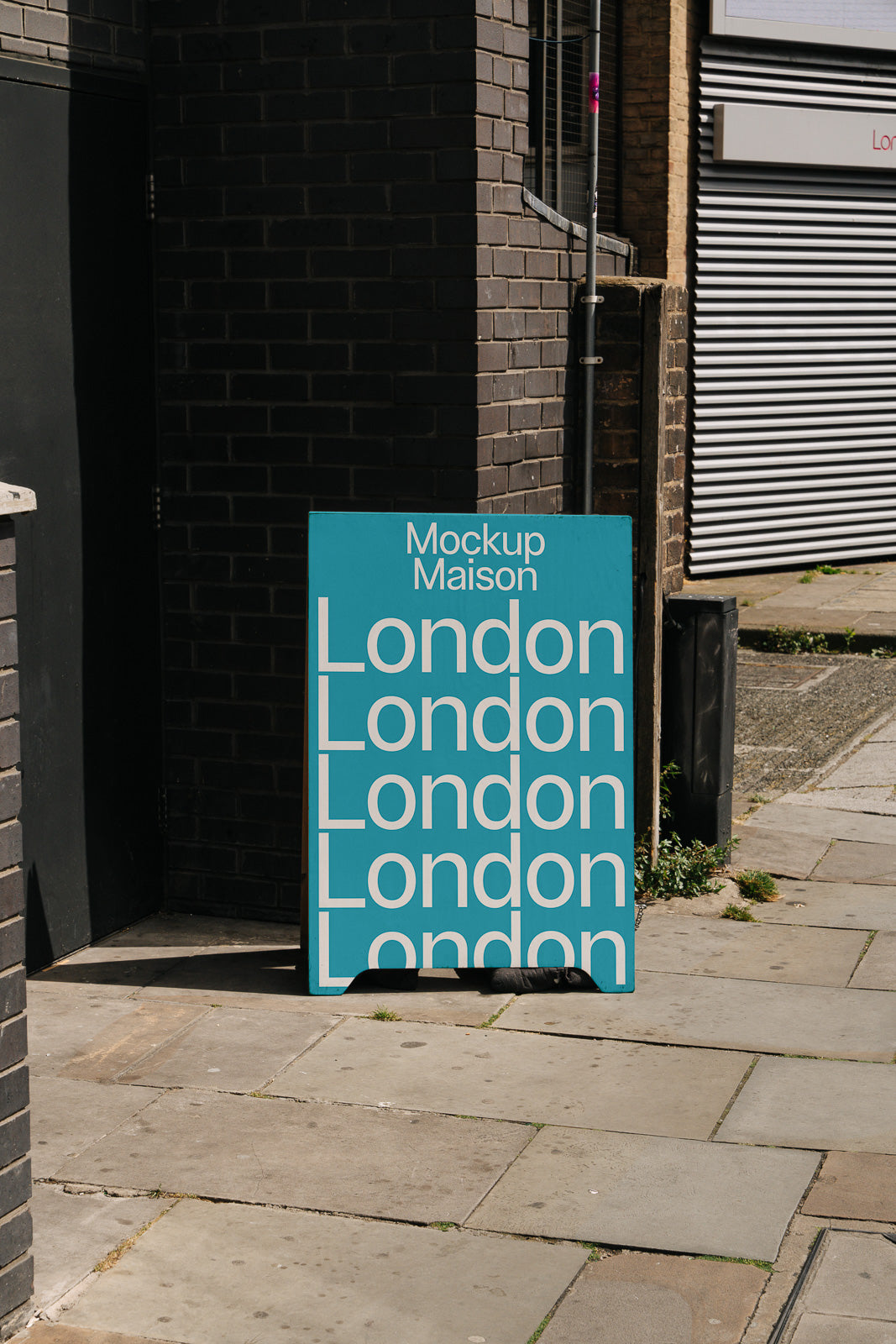 PSD mockup of a turquoise signboard with repeated "London" text, placed on an urban sidewalk near a dark brick wall and closed metal shutters.