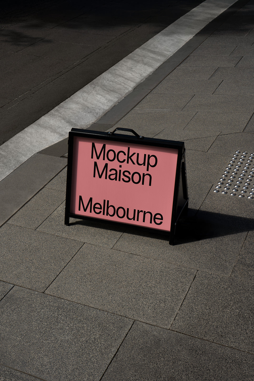 PSD mockup of a sidewalk sign displaying the text "Mockup Maison Melbourne" on a pink background, set against a stone pavement.