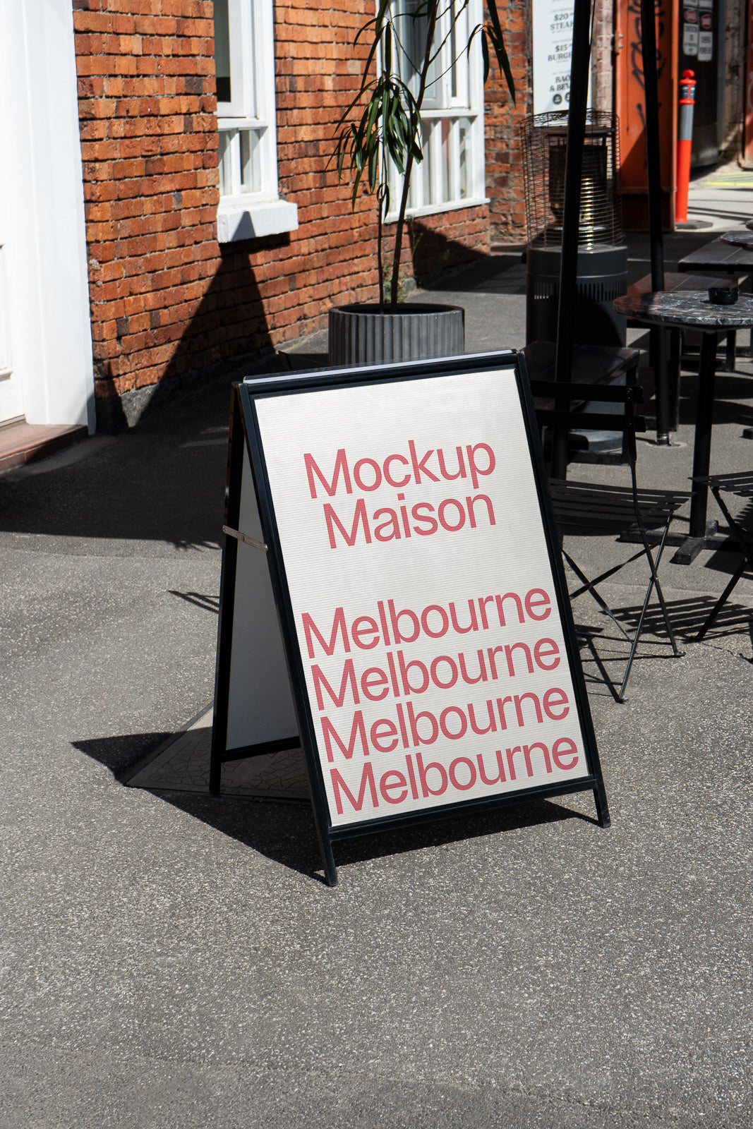 PSD mockup of a sidewalk A-frame sign displaying the text "Mockup Maison Melbourne" multiple times, set outdoors against a red brick wall.