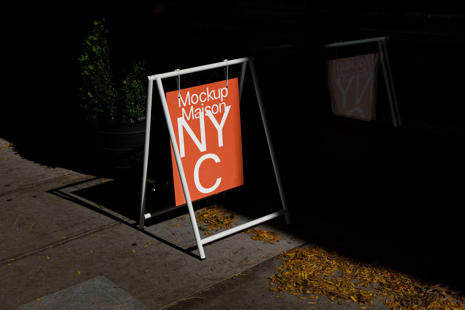 PSD mockup of a white A-frame sidewalk sign displaying an orange poster with "Mockup Maison NYC" text, set against a dark urban background.