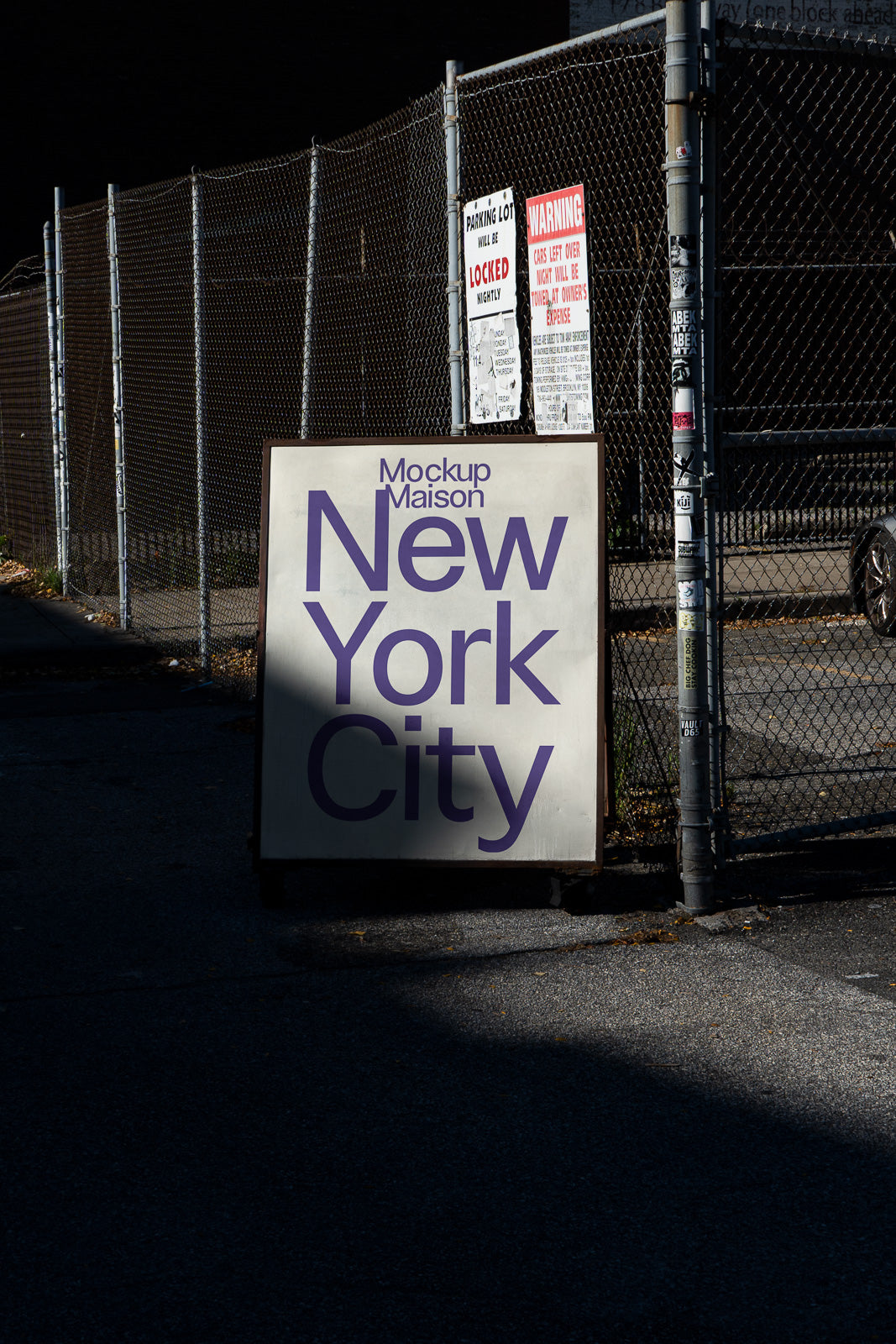PSD mockup of a signboard with "Mockup Maison New York City" in purple letters, placed on an urban street beside a chain-link fence with warning signs.