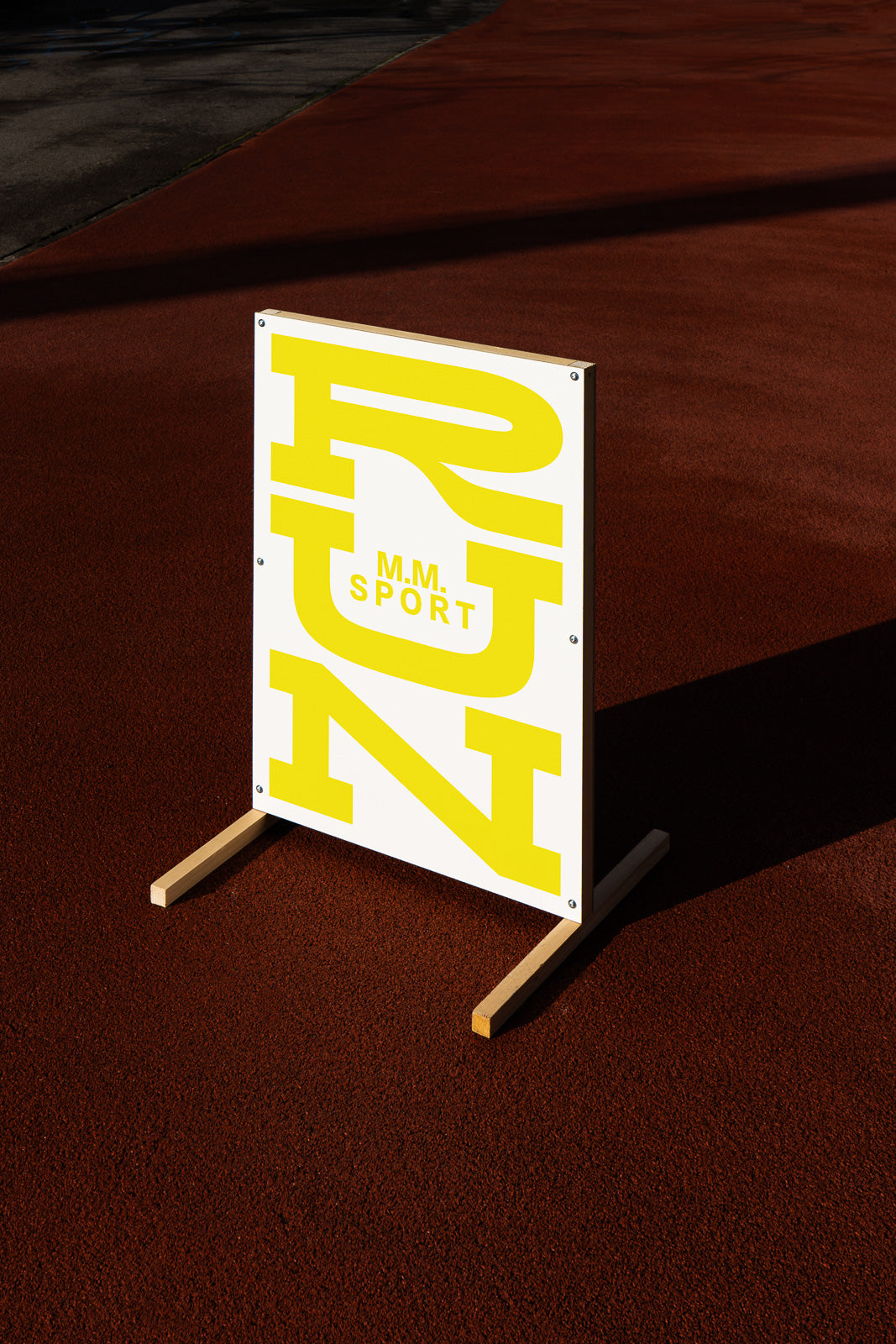 PSD mockup of a standing sign with yellow "RUN M.M. Sport" text on a white background, placed on a red track surface under sunlight.
