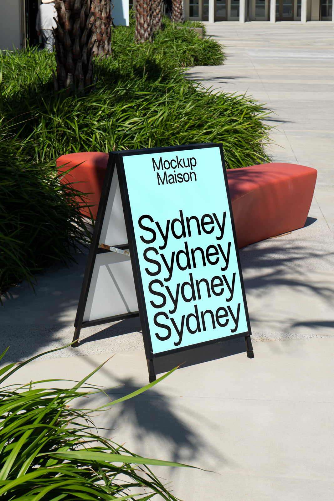 PSD mockup of a freestanding A-frame sign displaying repetitive text of "Sydney" in black on a light blue background, set outdoors beside red seating.