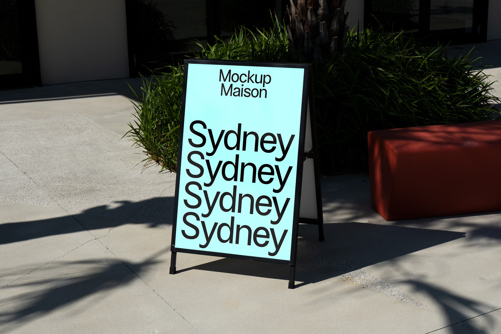 PSD mockup of an outdoor A-frame sign displaying "Sydney" repeatedly on a blue background, positioned on a sunny sidewalk near greenery and modern architecture.