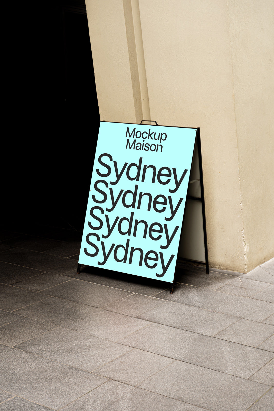 PSD mockup of an A-frame sidewalk sign displaying "Mockup Maison" and "Sydney" repeated in bold text against a pale blue background, leaning against a beige wall.