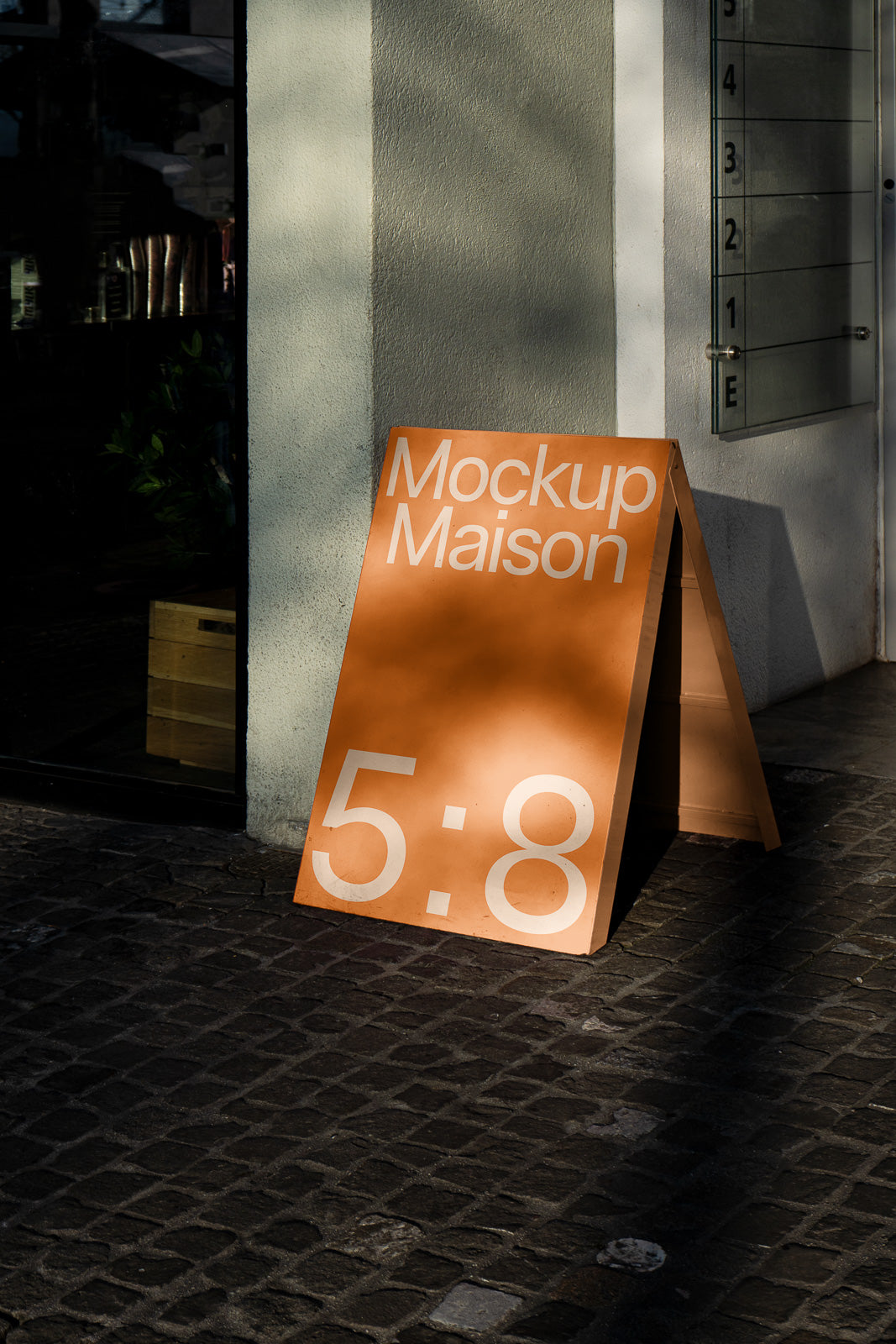 PSD mockup of an A-frame sidewalk sign with "Mockup Maison 5:8" displayed on a sunlit orange board leaning against a light-colored wall.