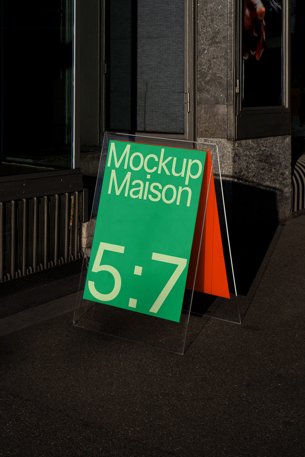 PSD mockup of an A-frame sign with green and orange panels displaying "Mockup Maison 5:7," positioned on a concrete sidewalk by a building entrance.