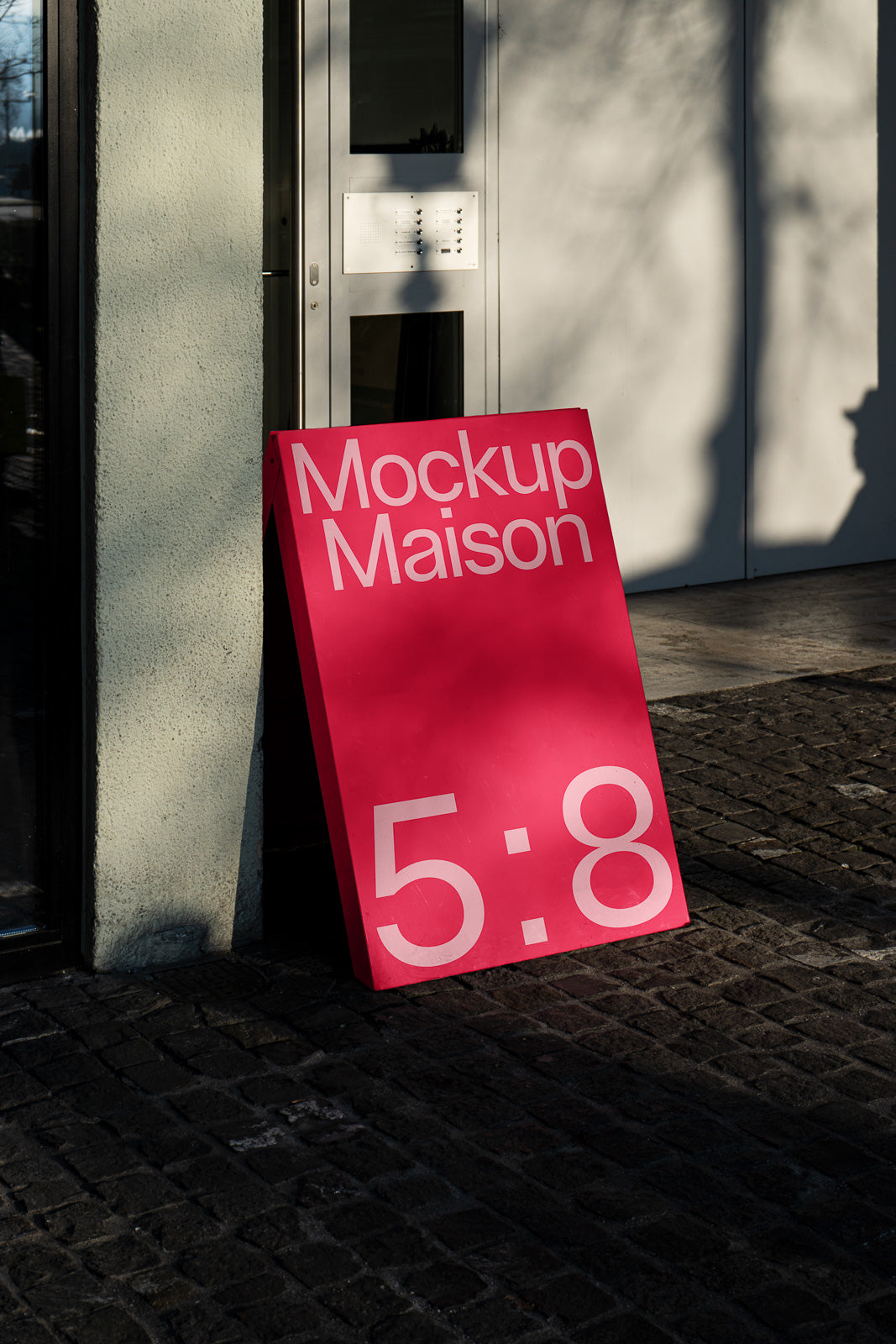 PSD mockup of a pink sandwich board with "Mockup Maison 5:8" in white text, placed on a cobblestone street near a building entrance.