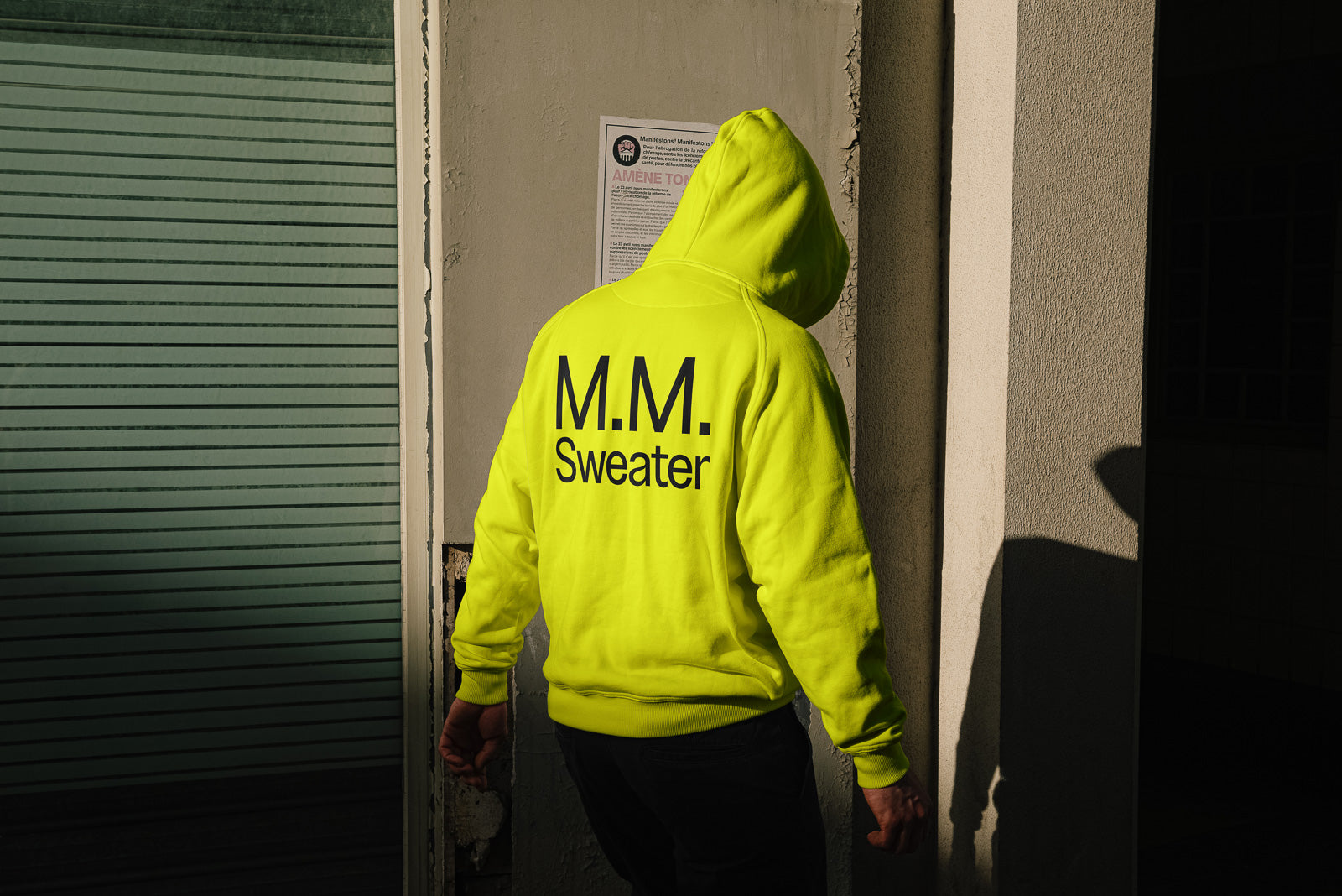 PSD mockup of a person wearing a bright yellow hoodie with "M.M. Sweater" printed on the back, standing outside near a building.