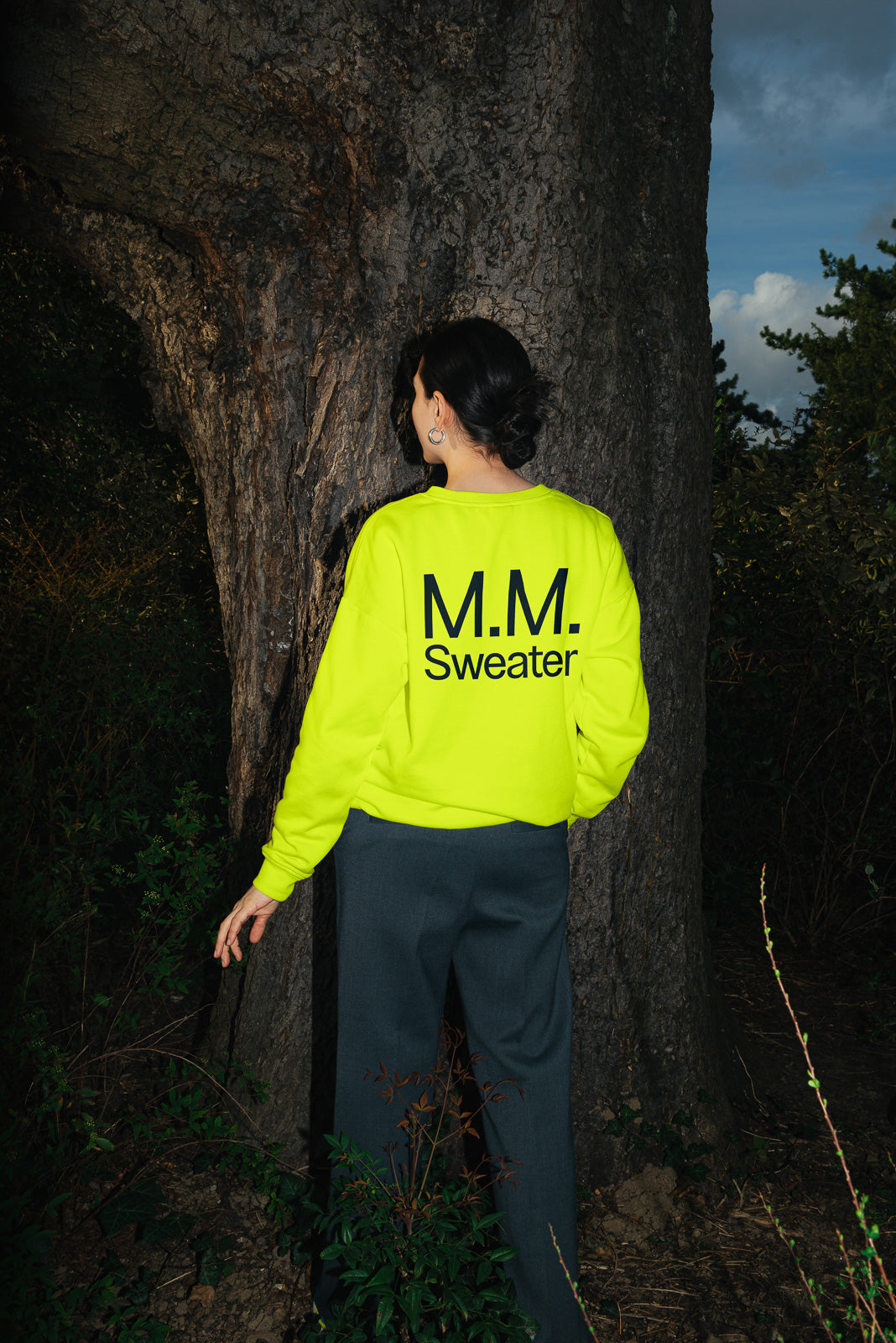 PSD mockup of person wearing a bright neon sweater with "M.M. Sweater" text, standing against a large tree trunk with surrounding foliage.