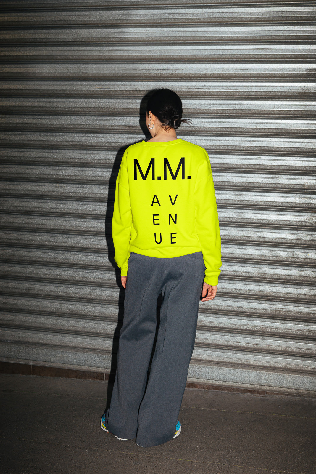PSD mockup of a person in a neon yellow sweatshirt with "M.M. AVENUE" on the back, standing in front of a metallic shutter.