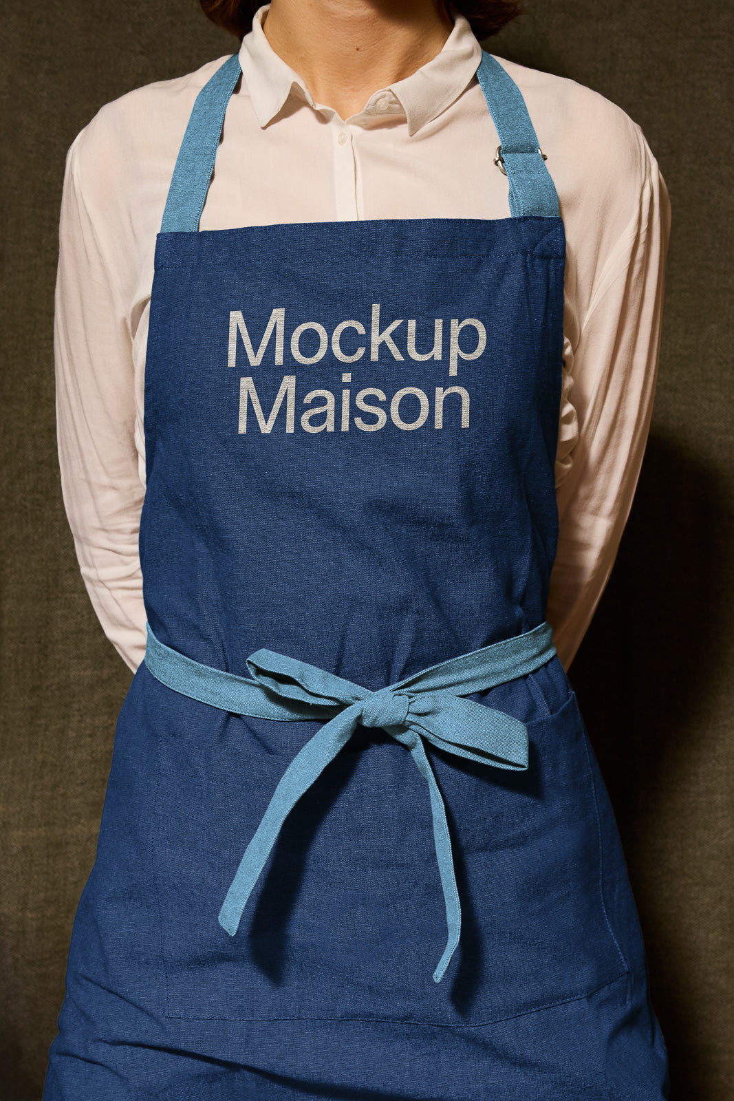 PSD mockup of a person wearing a blue apron with the words "Mockup Maison" printed on the front, tied at the waist over a white shirt.