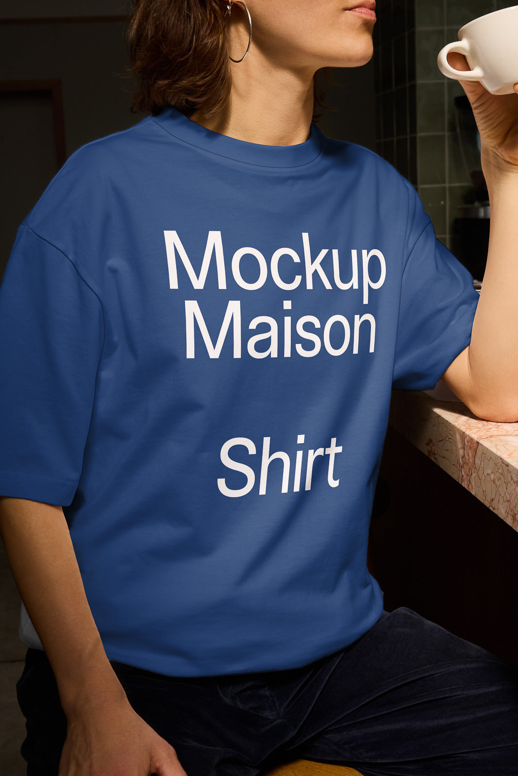 PSD mockup of a person wearing a blue t-shirt with the text "Mockup Maison Shirt" while sitting at a marble counter and holding a cup.