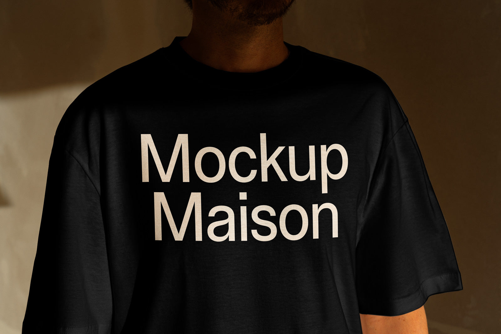 PSD mockup of a black T-shirt with "Mockup Maison" printed in large white letters, worn by an individual in partial shadow.