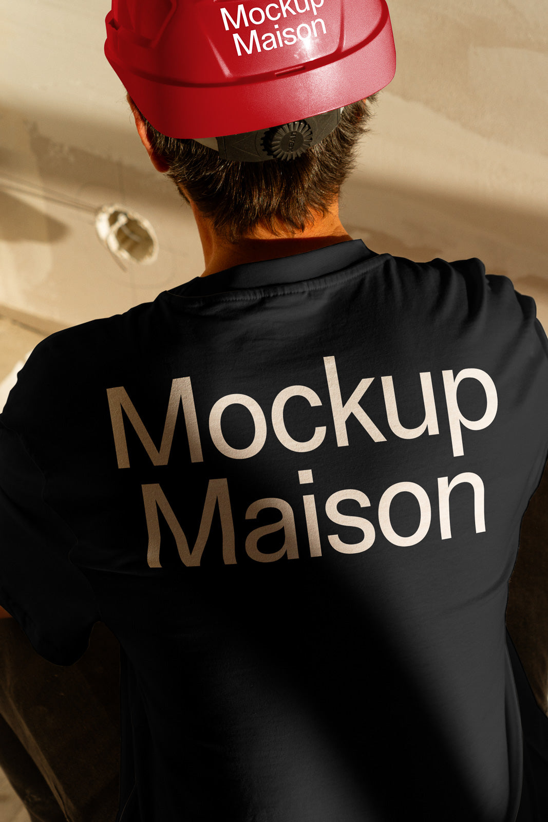 PSD mockup of a person in a black shirt with "Mockup Maison" printed on the back, wearing a red hard hat indoors.