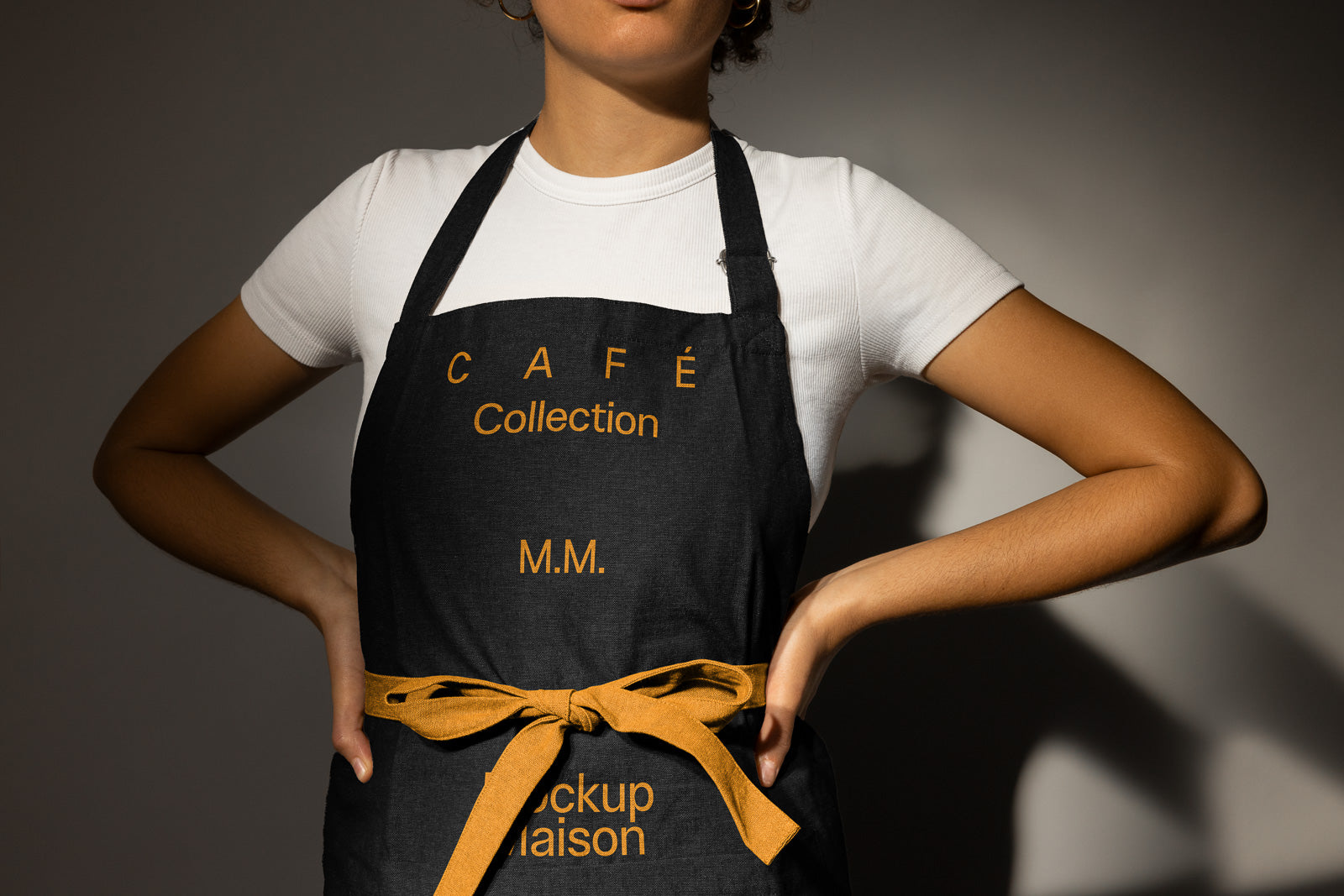PSD mockup of a person wearing a black apron with gold lettering, tied at the waist, over a white shirt, standing with hands on hips.