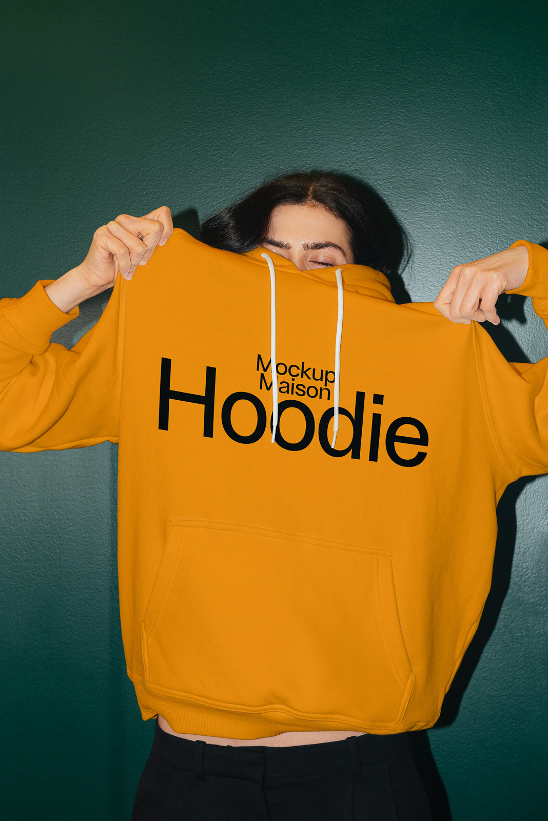 PSD mockup of a person holding up a yellow hoodie with "Mockup Maison Hoodie" printed in bold black letters, against a dark green background.