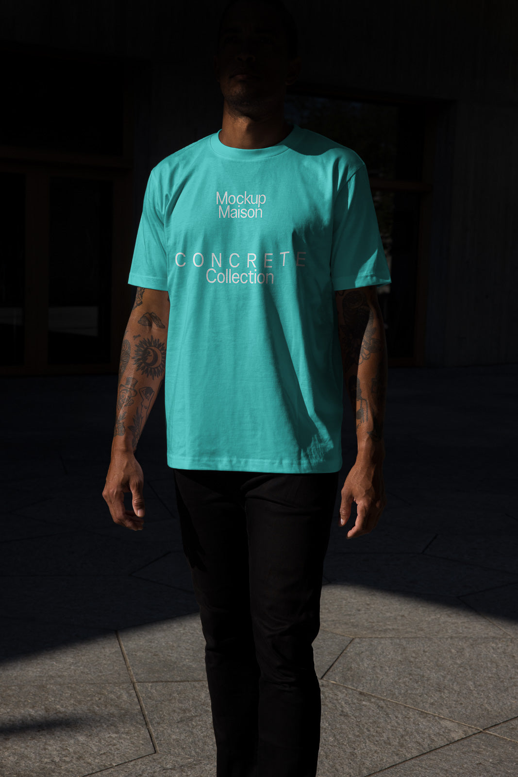 PSD mockup of a person in shadow wearing a light blue shirt with "Mockup Maison Concrete Collection" text, paired with black pants, standing on a stone surface.