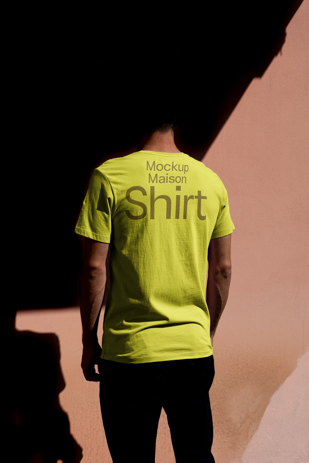 PSD mockup of a person standing against a pink wall, wearing a bright yellow T-shirt with the words "Mockup Maison Shirt" on the back.