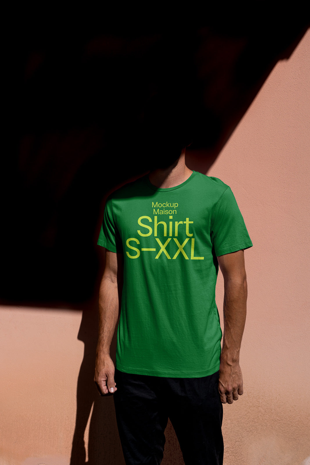PSD mockup of a person wearing a green t-shirt with "Mockup Maison Shirt S-XXL" text, standing against a peach-colored wall, partially in shadow.