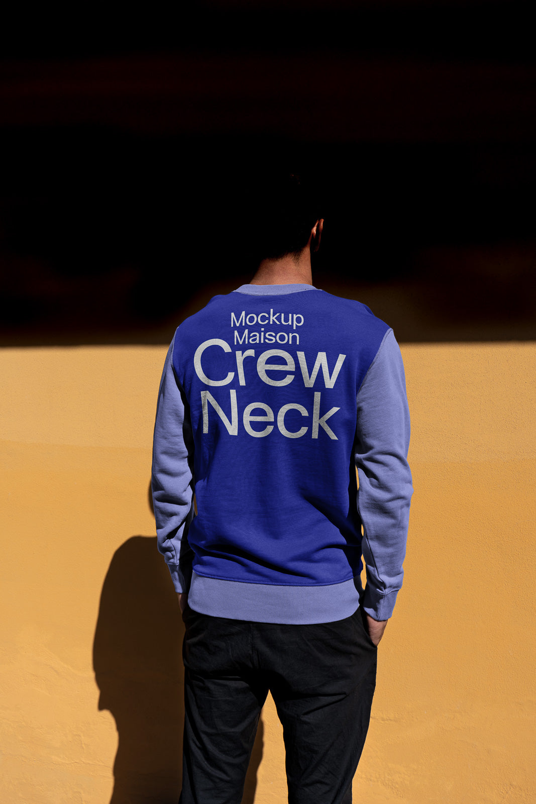 PSD mockup of a man wearing a blue crew neck sweatshirt, standing against a yellow wall, with "Mockup Maison Crew Neck" text on the back.