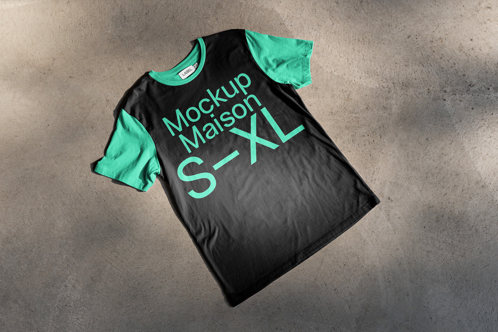 PSD mockup of a two-tone t-shirt featuring green sleeves and collar, with bold green text "Mockup Maison S-XL" on a black fabric, displayed on a concrete surface.