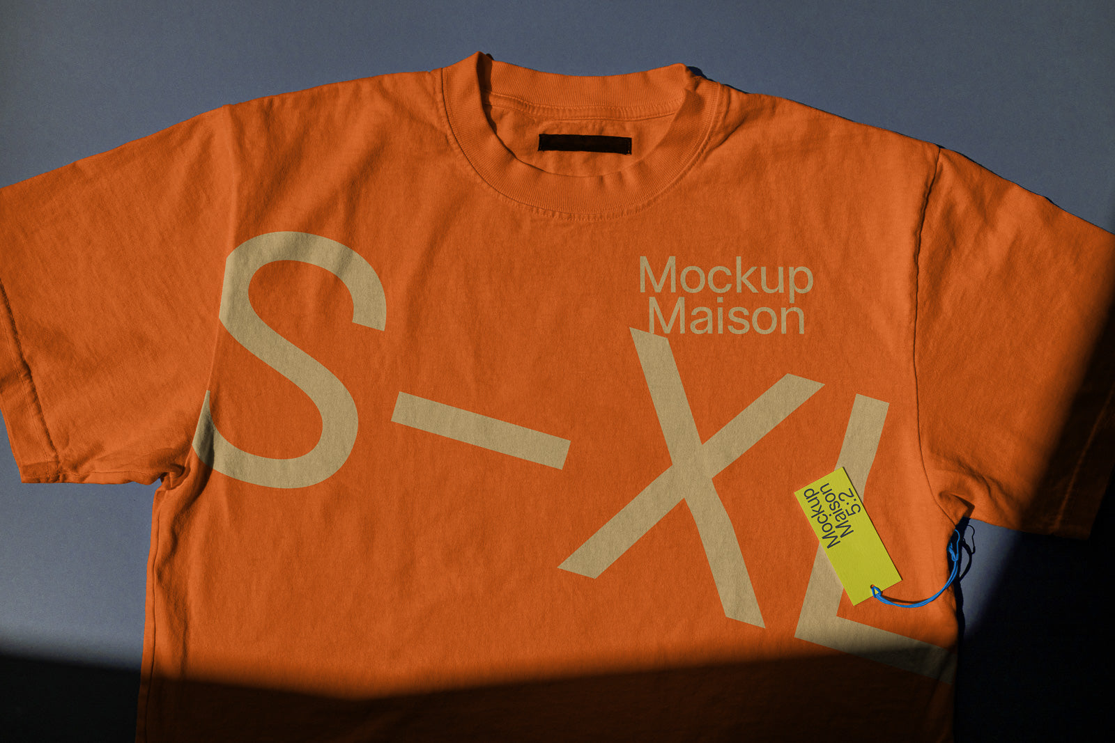 PSD mockup of an orange T-shirt with large lettering including "S-XL" and "Mockup Maison," featuring a tag attached with a blue string on a blue background.
