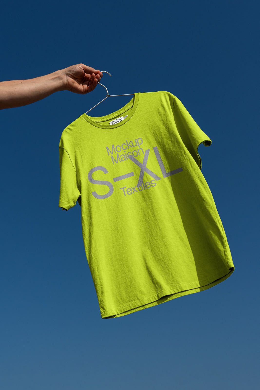 PSD mockup of a lime green T-shirt labeled "Mockup Maison S-XL Textiles" held on a hanger against a clear blue sky.