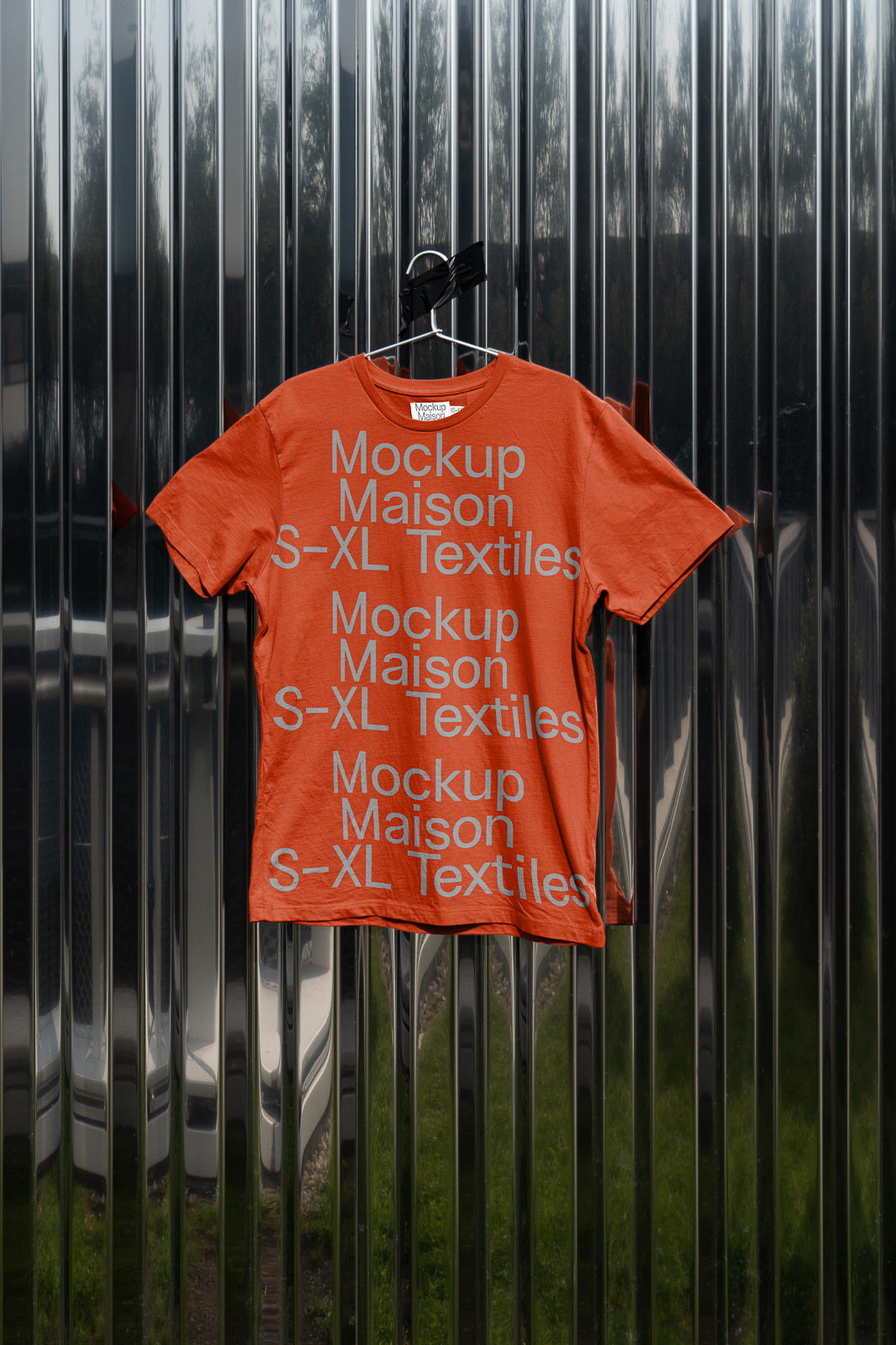 PSD mockup of an orange T-shirt with gray text reading "Mockup Maison S-XL Textiles" against a reflective, metallic background.