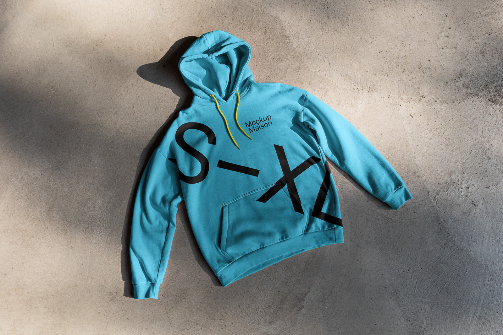 PSD mockup of a blue hoodie with black abstract letters, yellow drawstrings, and the text "Mockup Maison" printed on the chest, laid on a textured surface.