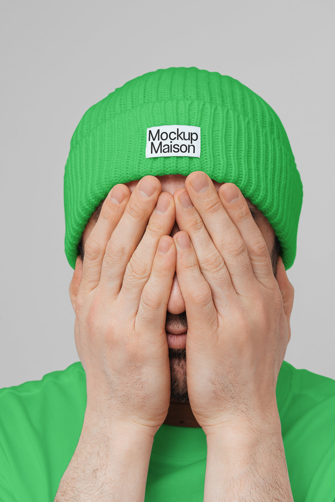 PSD mockup of a green beanie with "Mockup Maison" label, worn by a person covering their face with their hands against a neutral background.