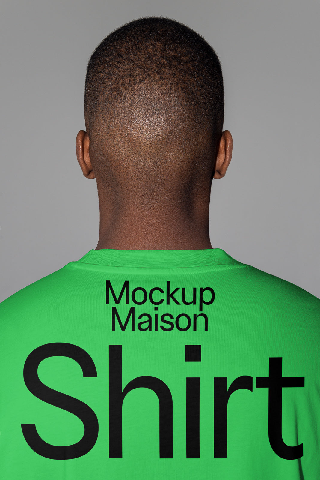 PSD mockup of a person in a green shirt with bold black text reading "Mockup Maison Shirt" on the back, viewed from behind against a gray background.