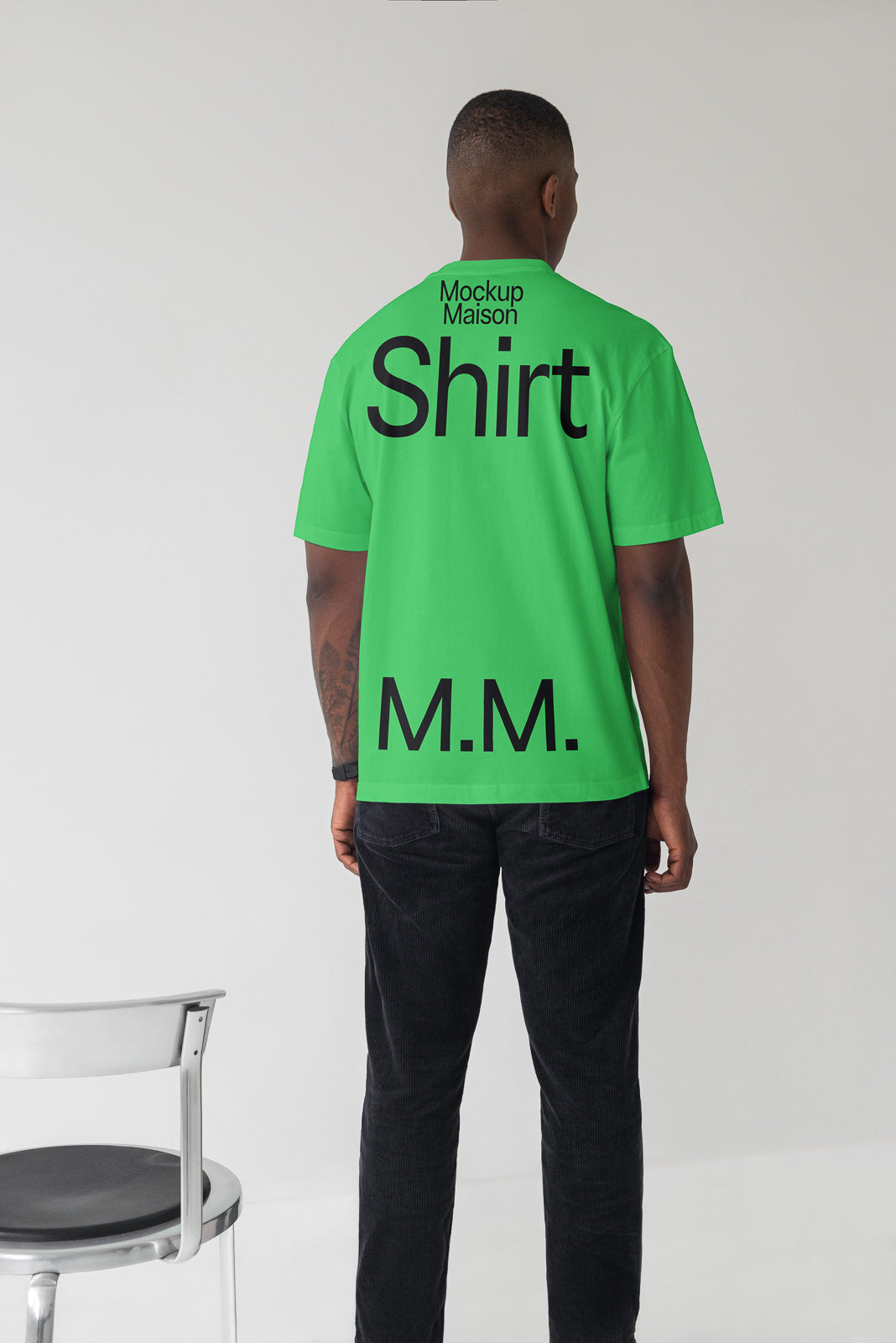 PSD mockup of a person wearing a green shirt with black text "Mockup Maison Shirt M.M." standing beside a metal chair.