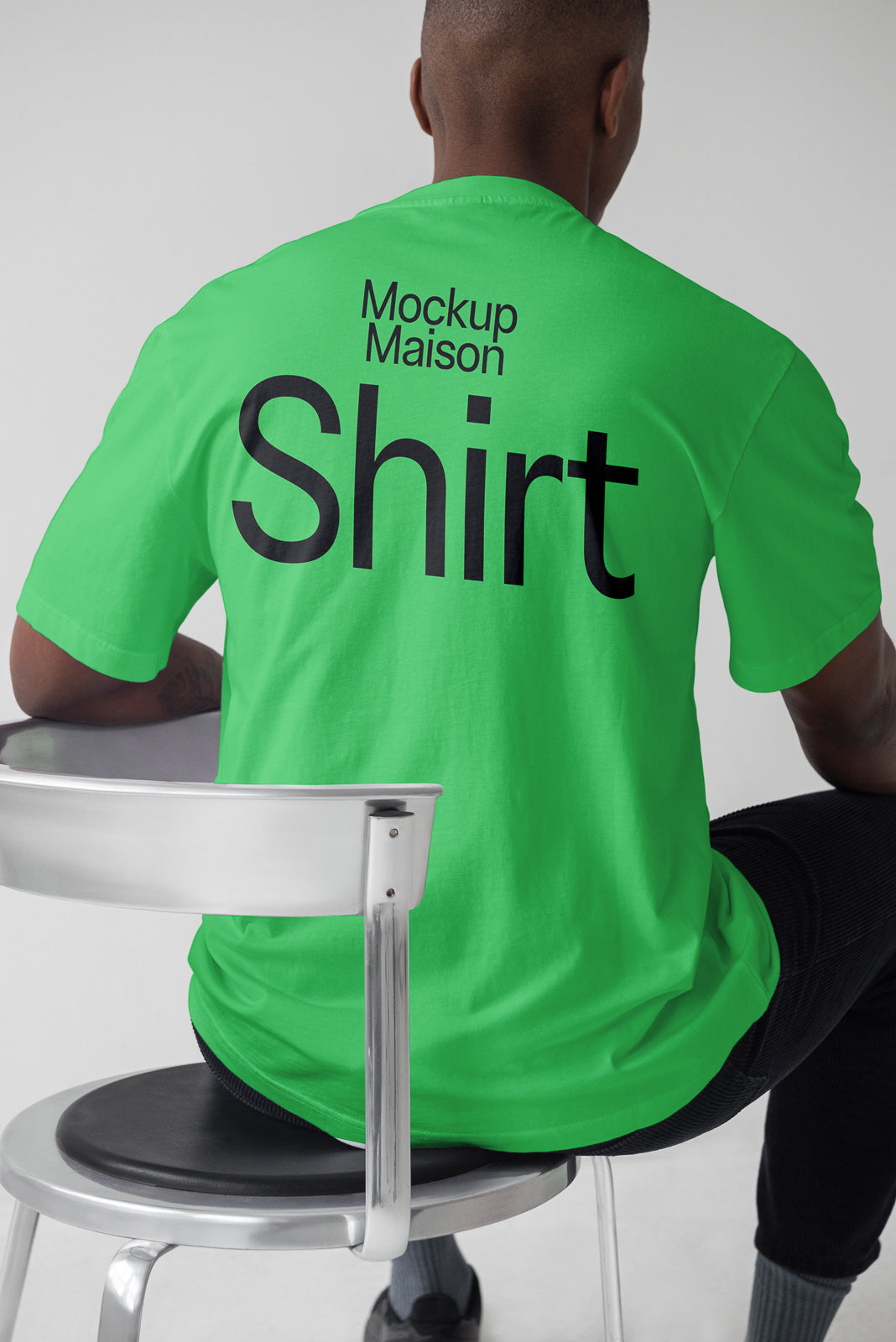 PSD mockup of a person sitting on a chair wearing a green T-shirt with the text "Mockup Maison Shirt" on the back, showing the shirt's design.