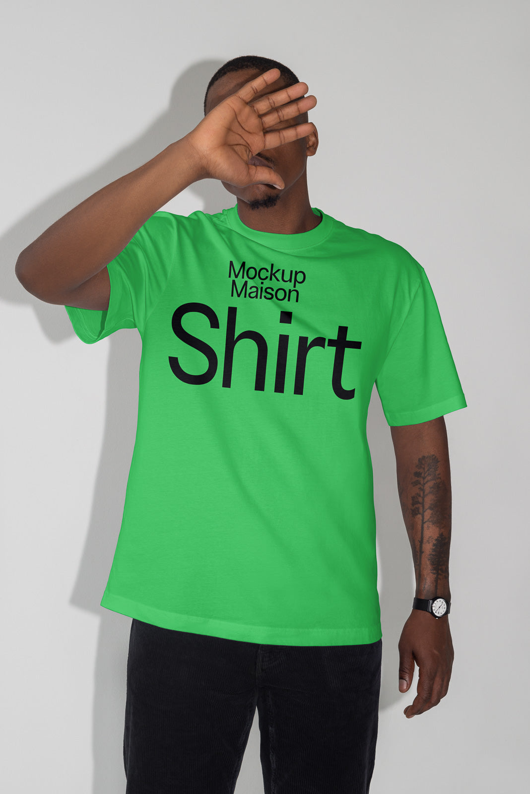 PSD mockup of a person wearing a bright green t-shirt with "Mockup Maison Shirt" text, hand raised to obscure face, tattoo visible on arm.