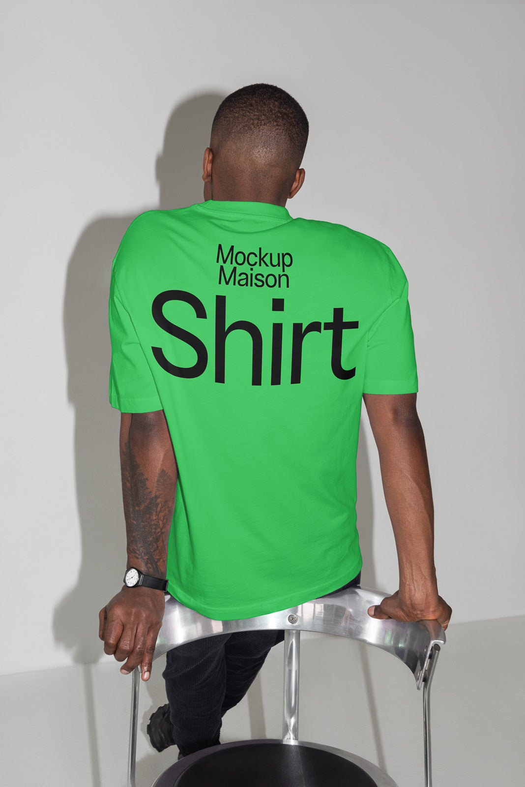 PSD mockup of a person wearing a vibrant green shirt with bold black text "Mockup Maison Shirt" on the back, seated on a metallic chair.