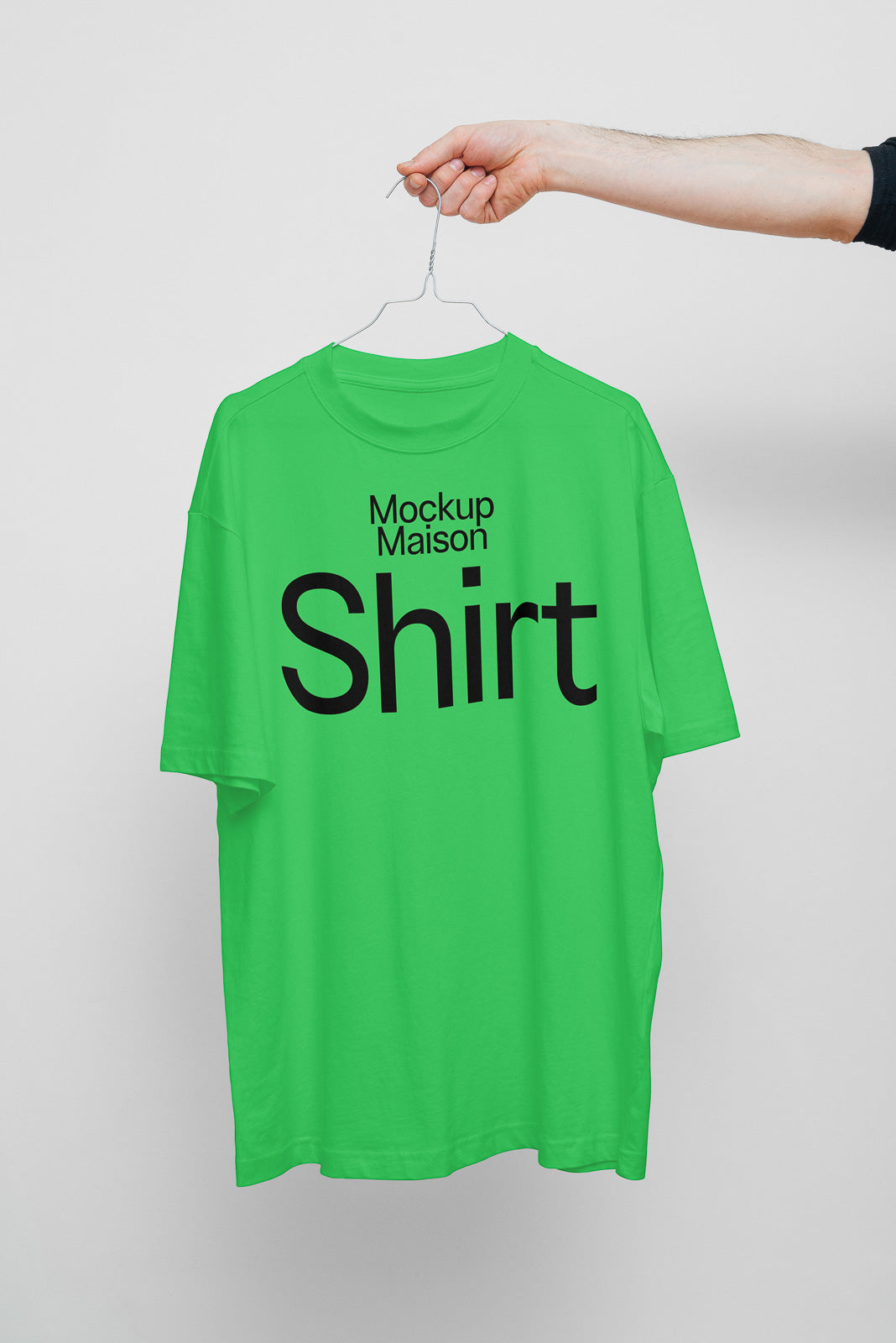 PSD mockup of a green T-shirt with the text "Mockup Maison Shirt," held on a hanger by a hand against a white background.