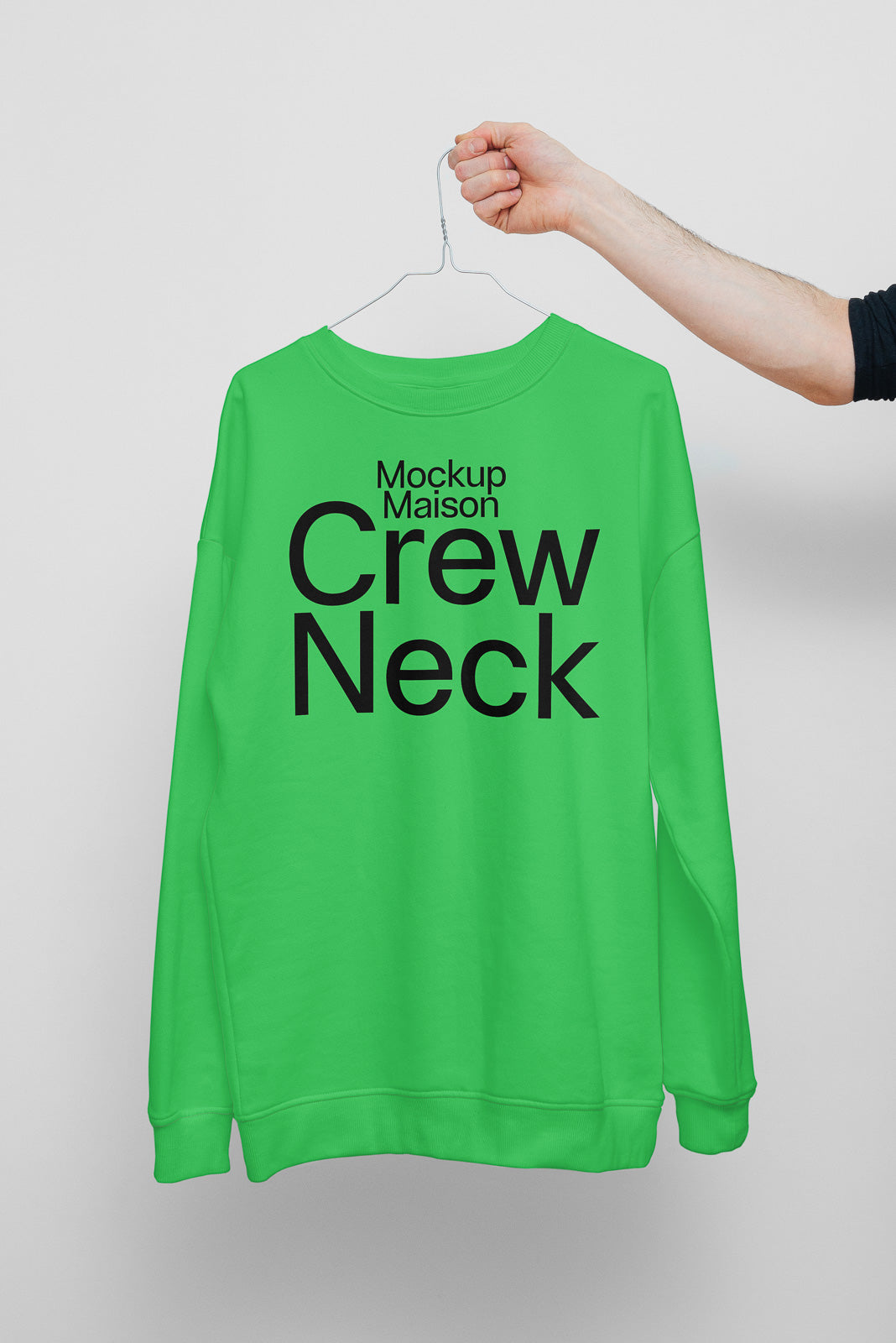 PSD mockup of a green crew neck sweatshirt on a hanger, held by a hand on a light gray background, displaying bold black text on the fabric.