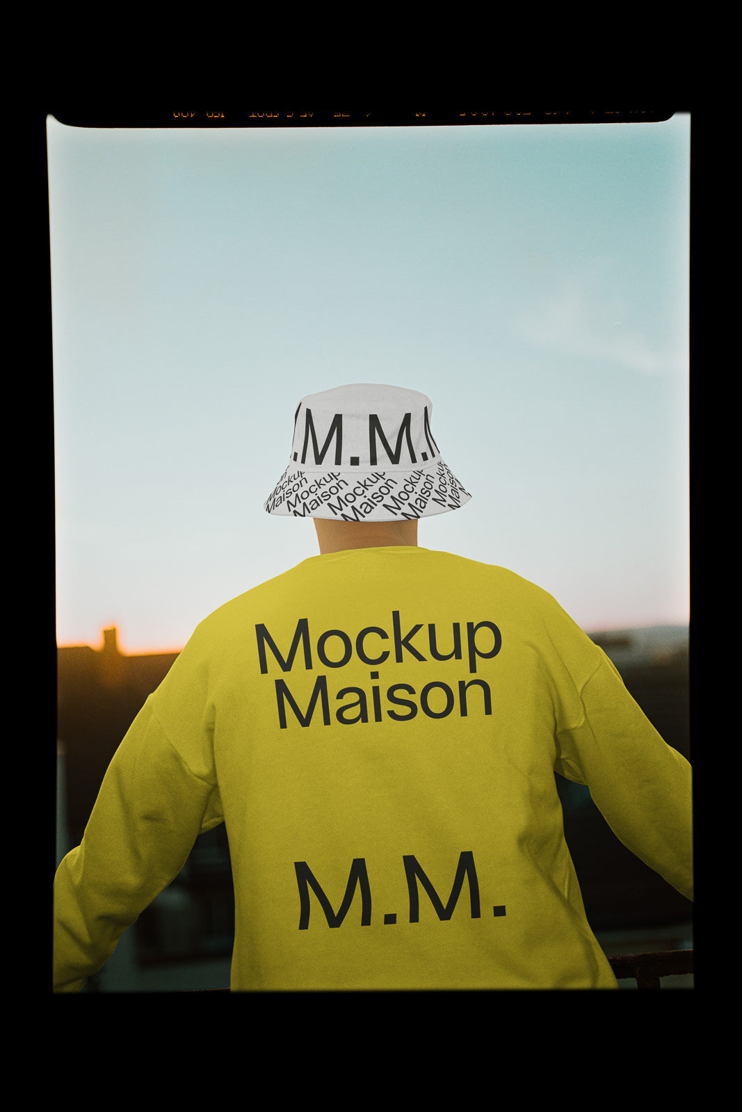 PSD mockup of a person wearing a yellow sweatshirt with "Mockup Maison" text and a bucket hat featuring "M.M." pattern against a sunset sky.