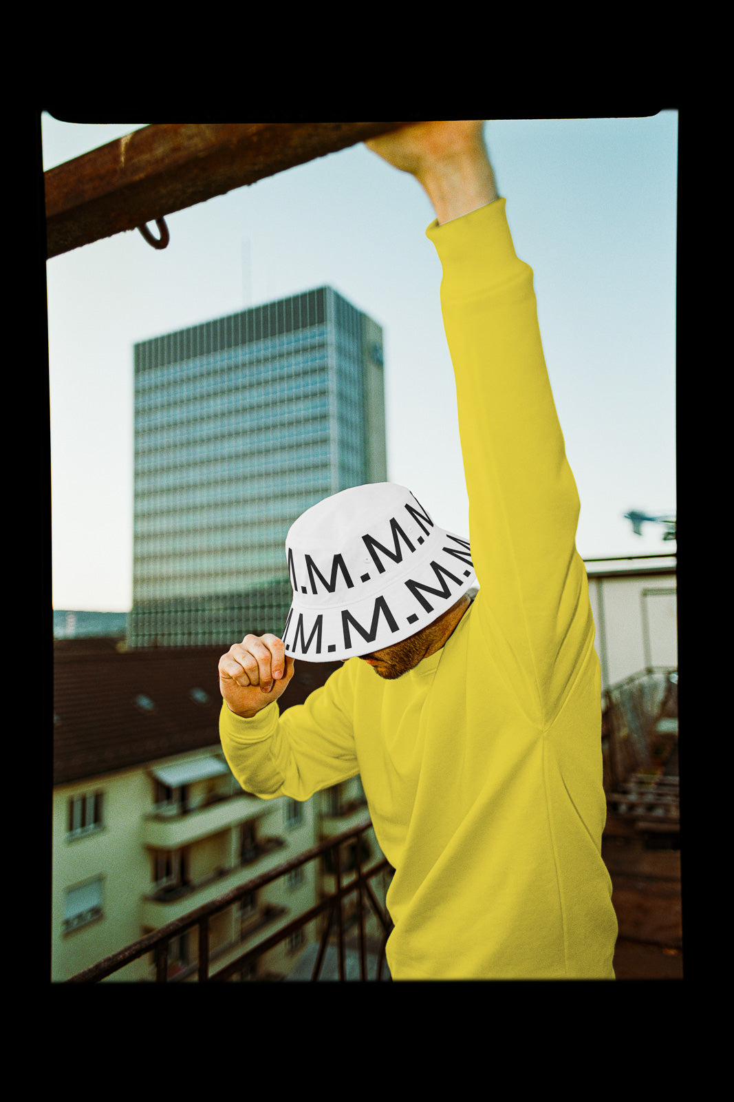 PSD mockup of a person wearing a white hat with bold lettering, paired with a yellow sweatshirt, set against an urban backdrop with a tall building.