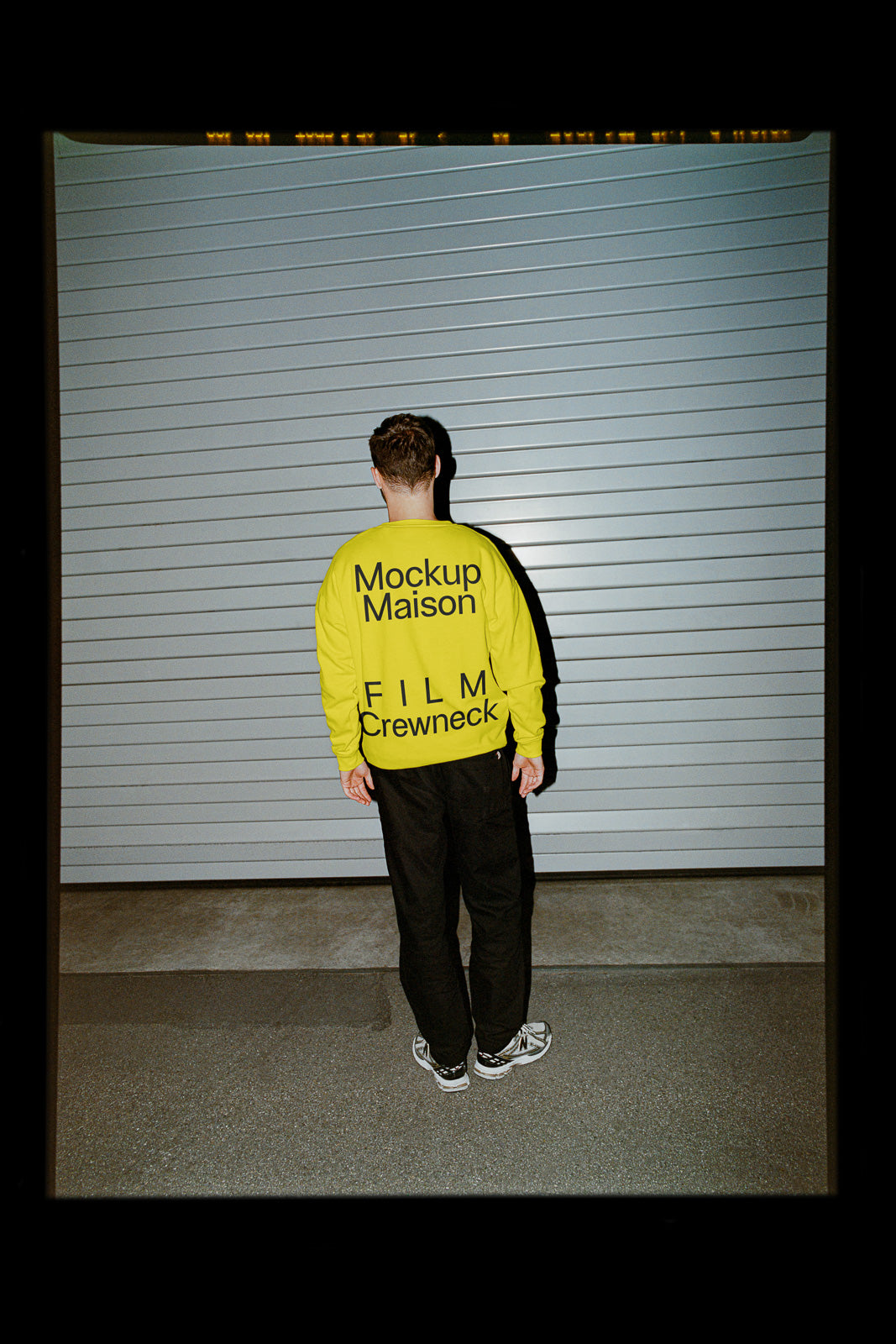 PSD mockup of person in yellow shirt with "Mockup Maison FILM Crewneck" text, standing in front of a gray corrugated metal wall.