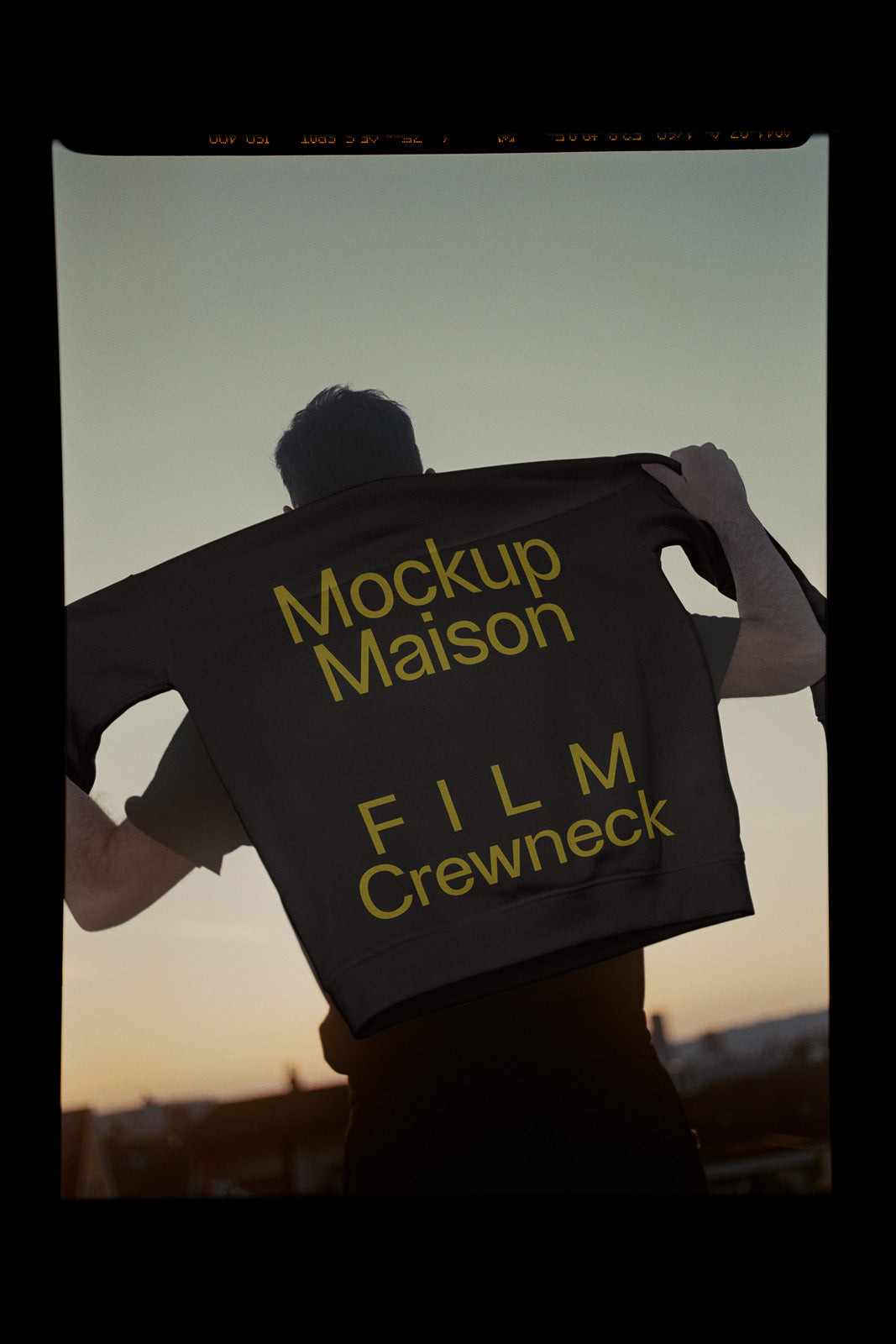 PSD mockup of a person holding a black sweatshirt with yellow text "Mockup Maison FILM Crewneck" against a sunset backdrop.