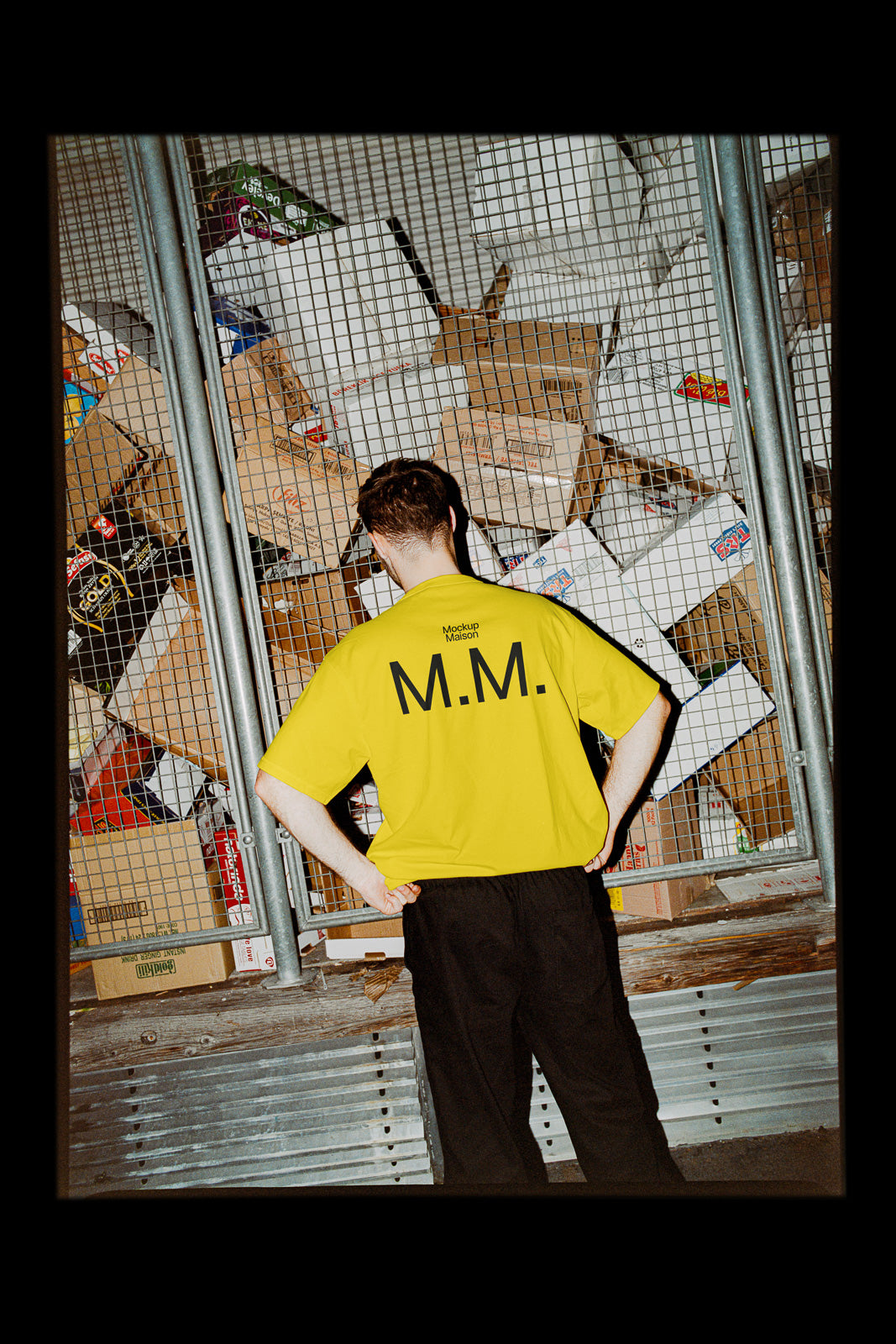 PSD mockup of a person in a bright yellow shirt marked "M.M." standing in front of a metal fence with cardboard boxes behind it.