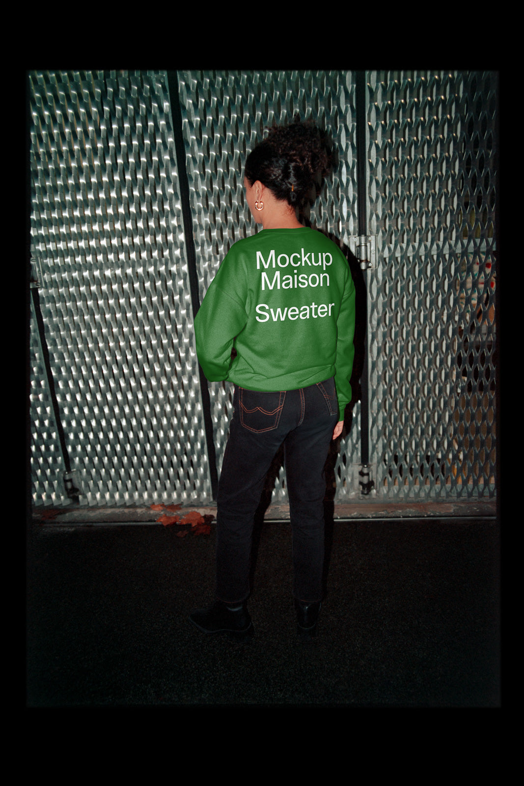 PSD mockup of a green sweater with "Mockup Maison Sweater" text, worn by a person standing against a textured metal fence.