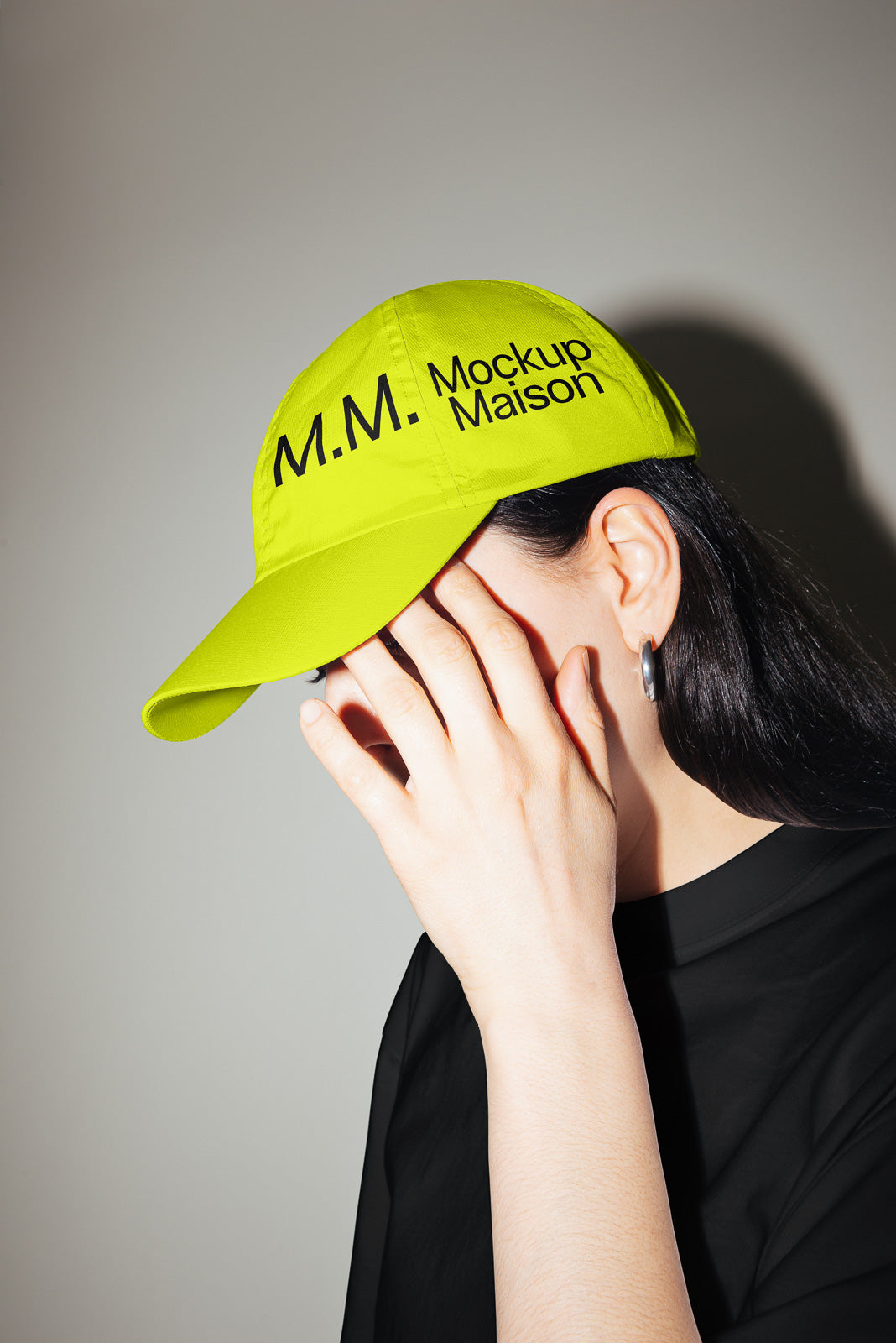 PSD mockup of a person wearing a bright yellow cap with "M.M. Mockup Maison" text, partially covering their face with their hand against a neutral background.