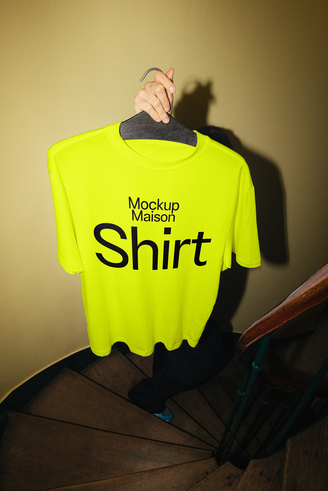 PSD mockup of a neon yellow T-shirt displaying the text "Mockup Maison Shirt," held on a hanger by a hand on a spiral staircase.